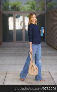 Full body positive stylish woman with thermos and bag closing eyes and smiling while walking on pavement outside modern building. Stylish female smiling and walking on pavement