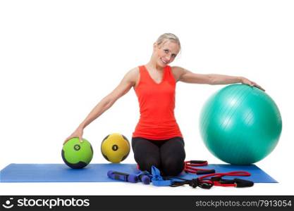 Full body shot of a smiling woman sitting on her knees on the mat and showing fitness equipment: dumb-bells,fitness ball, medicine balls and weights. Orange, green and black colors