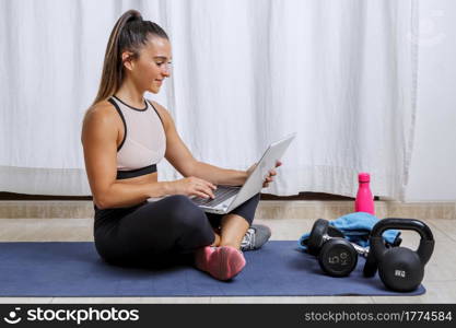 Full body side view of active young fit female in sportswear watching video tutorial on laptop while sitting on mat with sports equipment and preparing for workout at home. Positive woman using laptop during workout at home