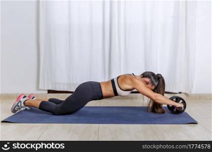 Full body side view of fit female doing knee roll out exercise with ab wheel while training core strength during workout at home. Fit woman training with ab roller