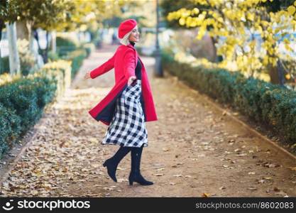 Full body side view of positive fema≤in red clothes sπnning around on path with fal≤n≤aves in park on autumn day. Cheerful woman sπnning around in park