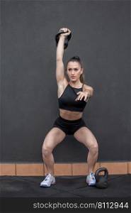 Full body strong female athlete lifting kettlebell over head and squatting against black wall during intense fitness workout in gym. Fit sportswoman squatting with kettlebell