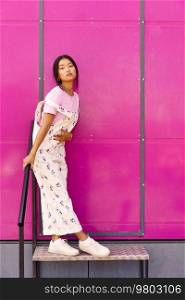 Full body stylish Asian female in casual clothes touching belly and looking at camera while leaning on banister near bright pink wall. Asian woman leaning on railing
