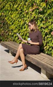 Full body stylish female manager in dress and high heeled shoes, with takeaway cup sitting on bench near bush and reading news on tablet during coffee break in park. Businesswoman reading data on tablet in park