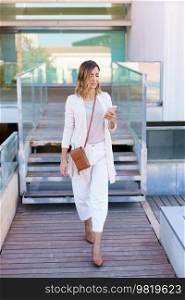 Full body woman in stylish clothes strolling on boardwalk and using mobile phone outside contemporary building. Female with smartphone walking on path