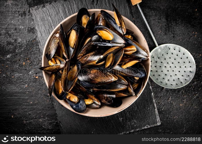 Full bowl with boiled mussels on a stone board. On a black background. High quality photo. Full bowl with boiled mussels on a stone board.