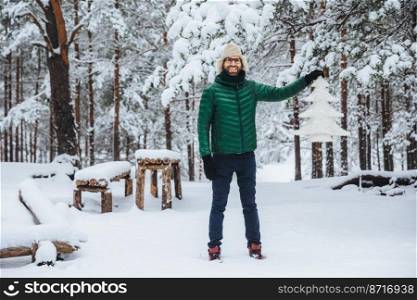 Full growth portrait of cheerful smiling male demonstrates white artificial fir tree, stands in winter forest has happy expression, glad to spend time outdoors. Winter and season concept