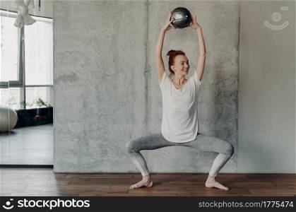 Full leghth shot of slim happy ginger female performing barre exercises with small fitball lifted up over head by two hands and sitting in deep plie in fitness studio, dressed in activewear. Slim pretty ginger woman enjoying barre exercises with small fitball