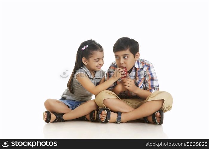 Full length girl feeding apple to brother while sitting over white background