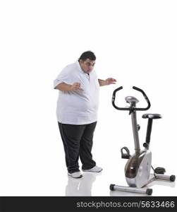 Full length of a shocked obese man looking at an exercise bike over white background