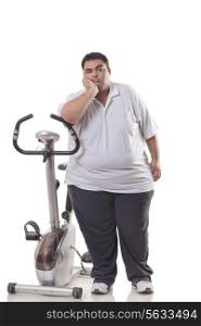 Full length of a tired obese man standing next to an exercise bike over white background