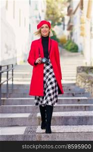 Full length of adult female with blond hair, in stylish outfit and beret smiling and looking at camera while walking on stairs on narrow street and photographing during holidays. Happy elegant lady walking downstairs on town street and smiling