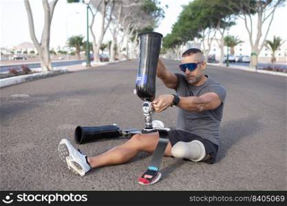 Full length of bearded male&utee in sunglasses and sportswear sitting on asphalt road and putting on artificial leg before training outside. Male runner putting on leg prosthesis before running