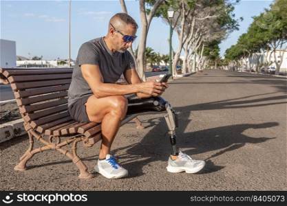 Full length of bearded male&utee with leg prosthesis sitting on bench resting after running while browsing mobile phone on sunny day. Male runner with disability using smartphone on street