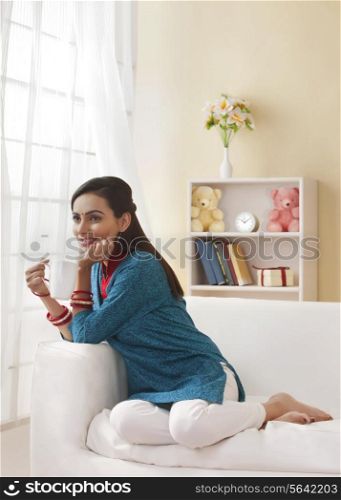 Full length of beautiful Indian woman having coffee on sofa
