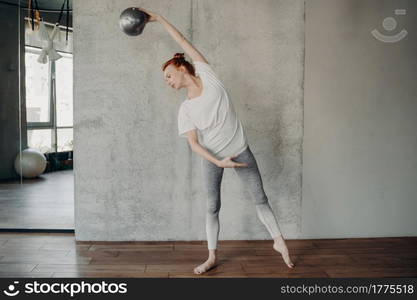 Full length of beautiful slender red haired lady standing barefoot in second position with fitball in hand raised over head in fitness studio against grey wall. Concept of barre workout and pilates. Red haired beautiful woman with fitball performing barre exercises