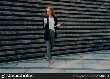 Full length of beautiful stylish red-haired business woman standing on city street outside with coffee cup, holding laptop, being on her way to office, female employee in spectacles going at work. Beautiful stylish red-haired business woman standing on city street outside with coffee cup