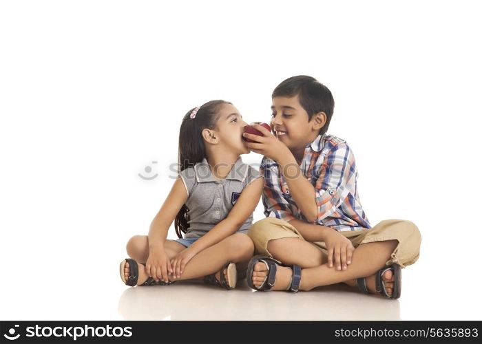 Full length of brother feeding apple to sister while sitting against white background