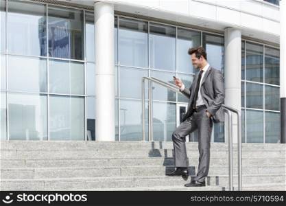 Full length of businessman text messaging through cell phone while standing on steps outside office