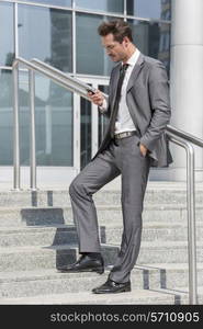 Full length of businessman text messaging through cell phone while standing on steps outside office