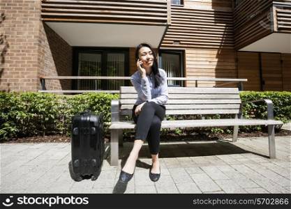Full length of businesswoman answering cell phone while sitting by luggage on bench against building