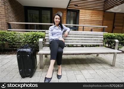 Full length of confident businesswoman sitting on bench by luggage against building
