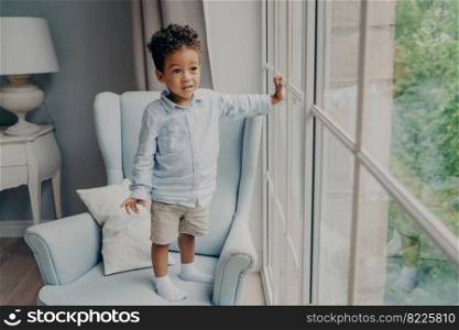 Full length of curious little afro american boy sweet toddler in stylish wear looking through window and waiting for mom while spending leisure time and playing alone at home in light room. Curious little afro american boy looking through window while standing on armchair at home