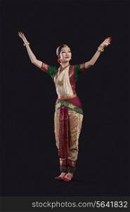 Full length of dancer with arms raised performing Bharatanatyam against black background