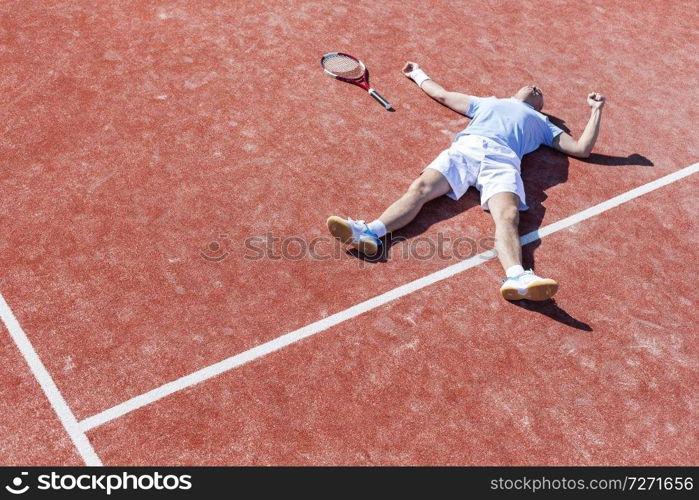 Full length of disappointed mature man lying by tennis racket on court during summer