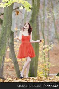 Full length of fashionable young woman in vibrant red dress relaxing in park. Red hair happy girl playing with colorful leaves outdoor. Autumnal scenery. Fashion and happiness.