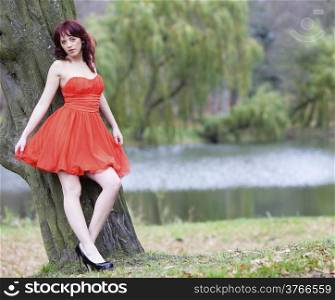 Full length of fashionable young woman in vibrant red dress relaxing in park. Red hair girl posing outdoor. Autumnal scenery. Fashion.