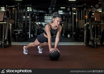 Full length of focused strong female athlete with long dark hair in sportswear and sneakers doing plank with medicine ball and lookign away during workout in gym. Serious fit sportswoman doing medicine ball plank exercise in gym