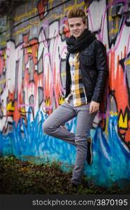 Full length of handsome trendy man outdoor in city setting, male model wearing black jacket scarf and checked shirt against colorful graffiti wall