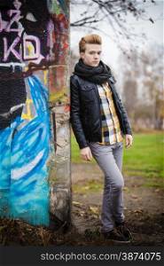 Full length of handsome trendy man outdoor in city setting, male model wearing black jacket scarf and checked shirt against colorful graffiti wall