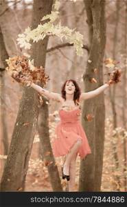 Full length of happy woman in red dress doing fun playing with leaves in autumnal park or forest