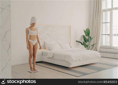 Full length of healthy young slender woman in underwear with bath towel wrapped on head after morning shower looking down while standing on scales if she has lost weight in modern bedroom at home. Shot of healthy young slender woman in underwear standing on scales in bedroom