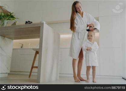 Full length of loving mother in white bathrobe brushing hair of cute little daughter after taking shower or bath while standing in modern kitchen at home, mom teaching small child good hygiene habits. Loving mom brushing hair of cute little daughter after taking bath at home