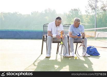 Full length of mature male friends talking while sitting with tennis rackets and balls on chair at court
