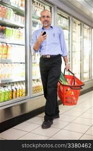 Full length of mature man in shopping store carrying basket and using mobile phone while walking