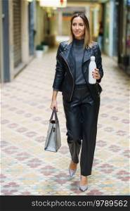 Full length of modern businesswoman in leather pants and jacket wearing heels and holding reusable bottle with drink and handbag walking in alley. Confident stylish lady with eco friendly bottle in town