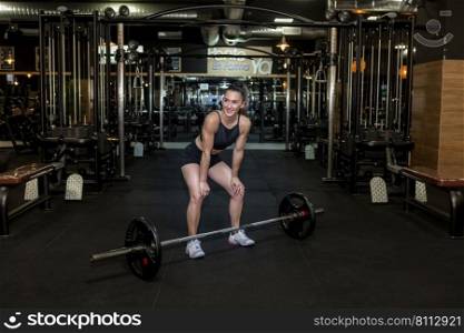 Full length of positive young fit female athlete with long hair in activewear leaning forward and smiling while resting during workout with heavy barbell in modern gym. Muscular lady smiling during training with barbell in sports club