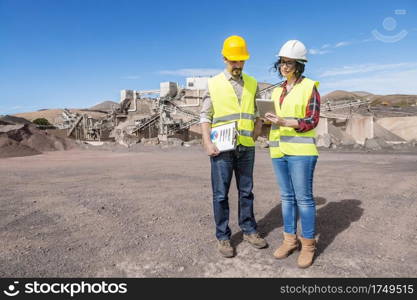 Full length of professional male and female engineers in hardhats and waistcoats using tablet and discussing engineering process while standing on industrial construction site. Construction colleagues with tablet discussing draft of industrial facility