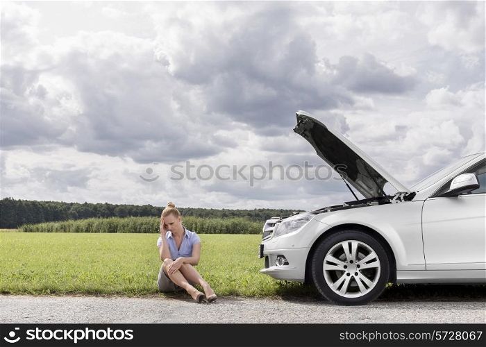Full length of sad businesswoman sitting by broken down car at countryside