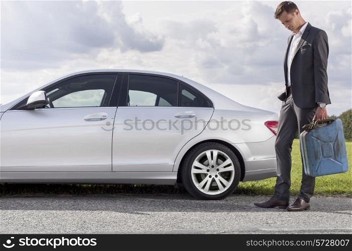 Full length of sad young businessman carrying gas can by broken down car at countryside