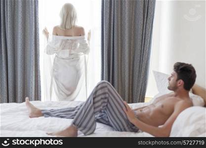 Full length of shirtless young man looking at woman standing by hotel window