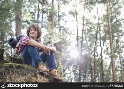 Full length of smiling male hiker looking away while sitting on cliff in forest