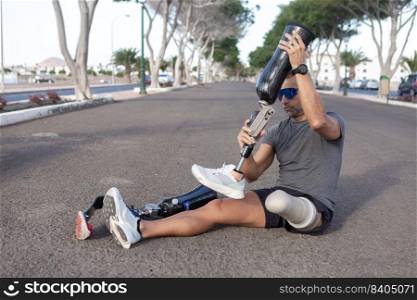 Full length of sportive male&utee in activewear sitting on asphalt road and taking off artificial leg while training in city. Sportsman changing leg prosthesis on road