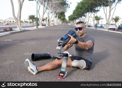 Full length of sportsman with disability sitting on road and putting on blade leg prosthesis before running training in city park. Male&utee sitting on road and putting on artificial leg