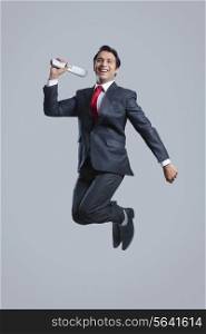 Full length of successful businessman with newspaper jumping over gray background