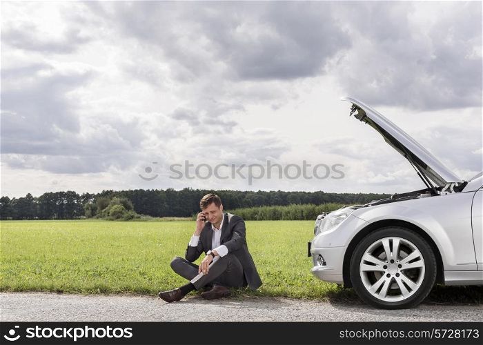 Full length of unhappy young businessman using cell phone by broken down car at countryside